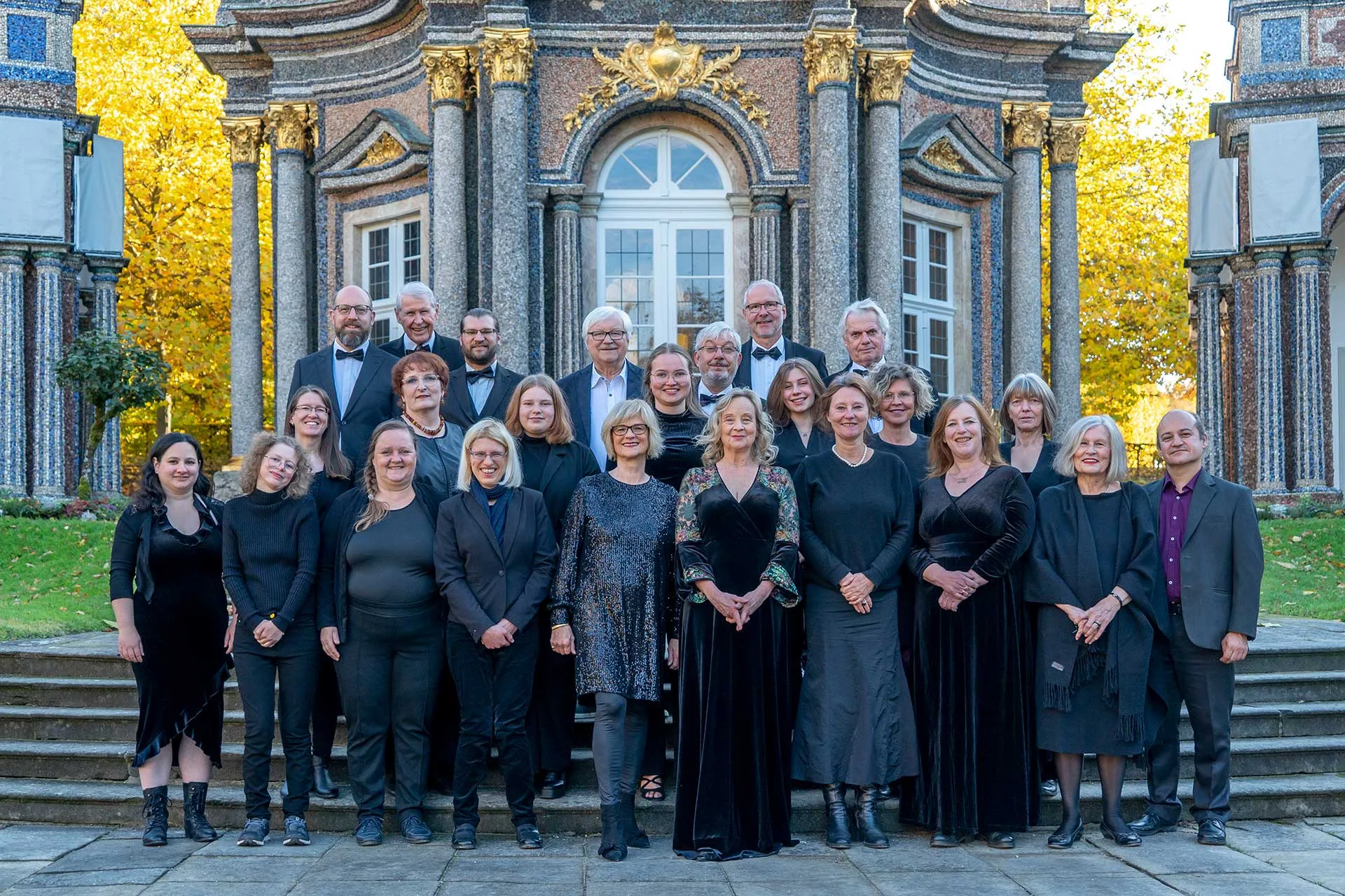 Der Zamirchor in festlicher Konzertkleidung auf den Stufen vor dem Sonnentempel der Eremitage in Bayreuth, im Hintergrund Herbstlaub