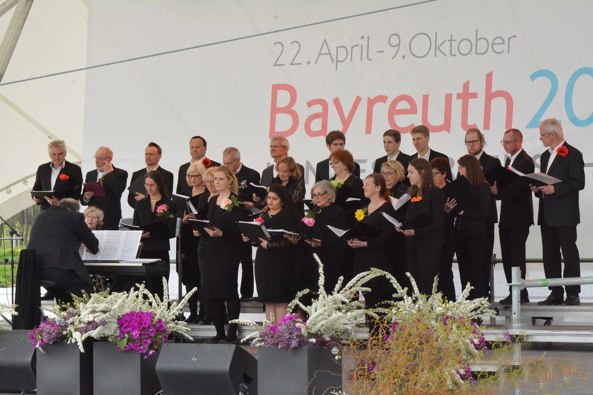 Der Zamirchor singt auf einer Freilichtbühne in Bayreuth, vorne Blumenschmuck und der Pianist am Keyboard